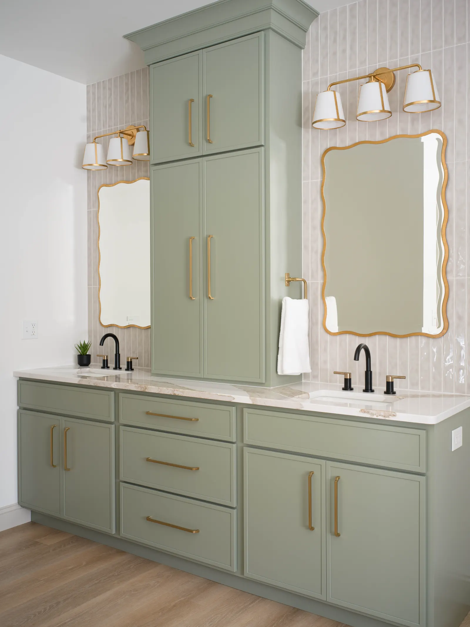 Sage green double vanity with stone tops, a tall centre cabinet between the sinks and gold-framed mirrors